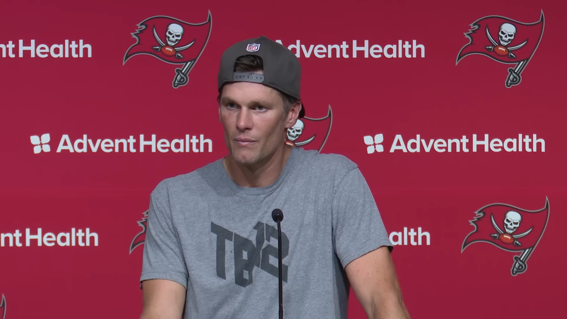 Tom Brady wearing a gray t-shirt and a backward baseball cap, speaking at a press conference with a red backdrop featuring the Tampa Bay Buccaneers logo and