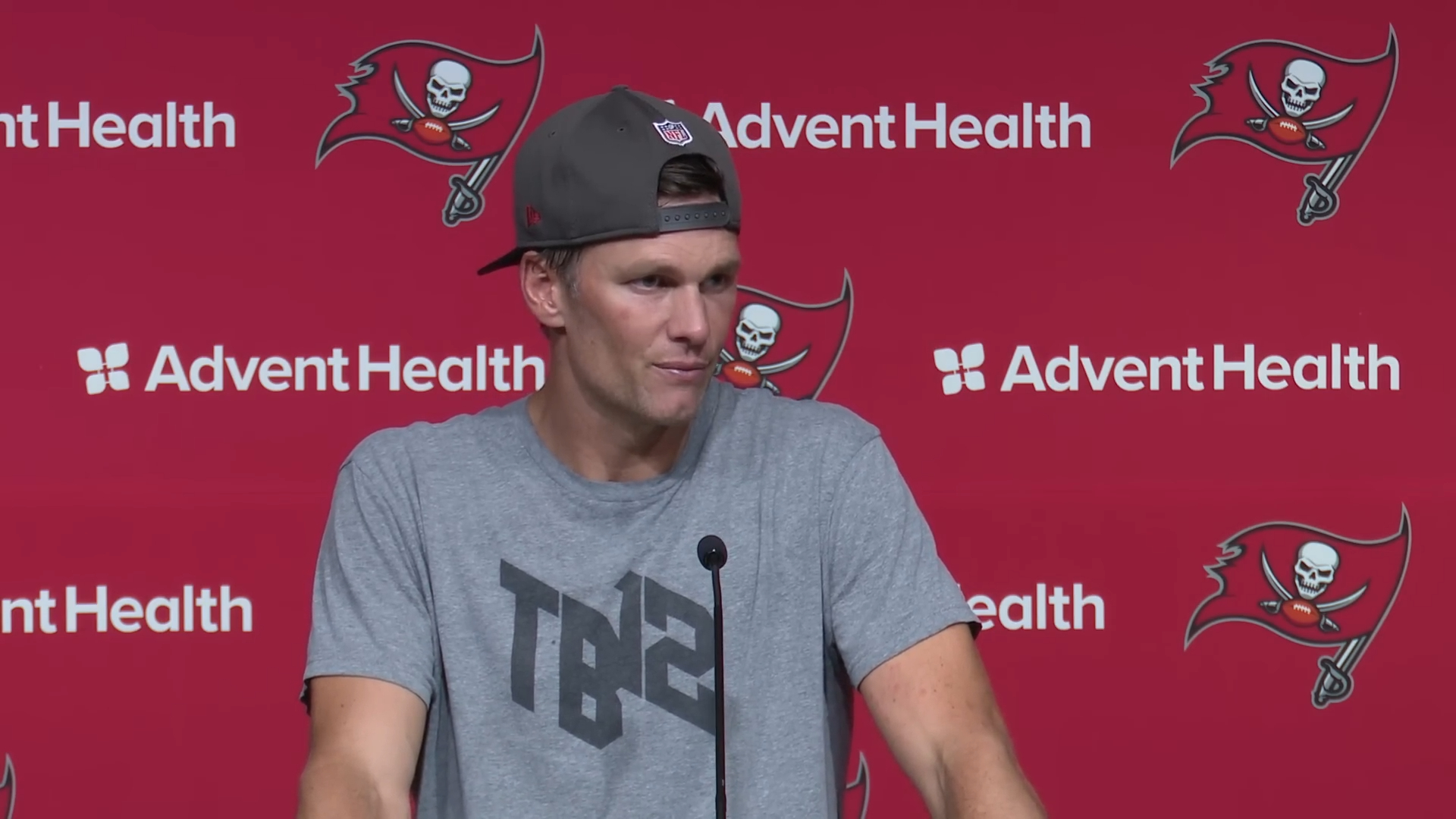 Tom Brady at a press conference with a red AdventHealth background.