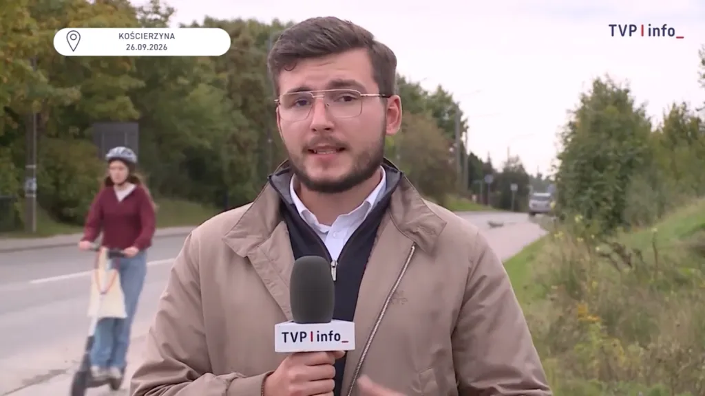 A male TVP Info reporter stands outdoors with a microphone, while a teenage girl on a scooter is visible in the background just before an accident.