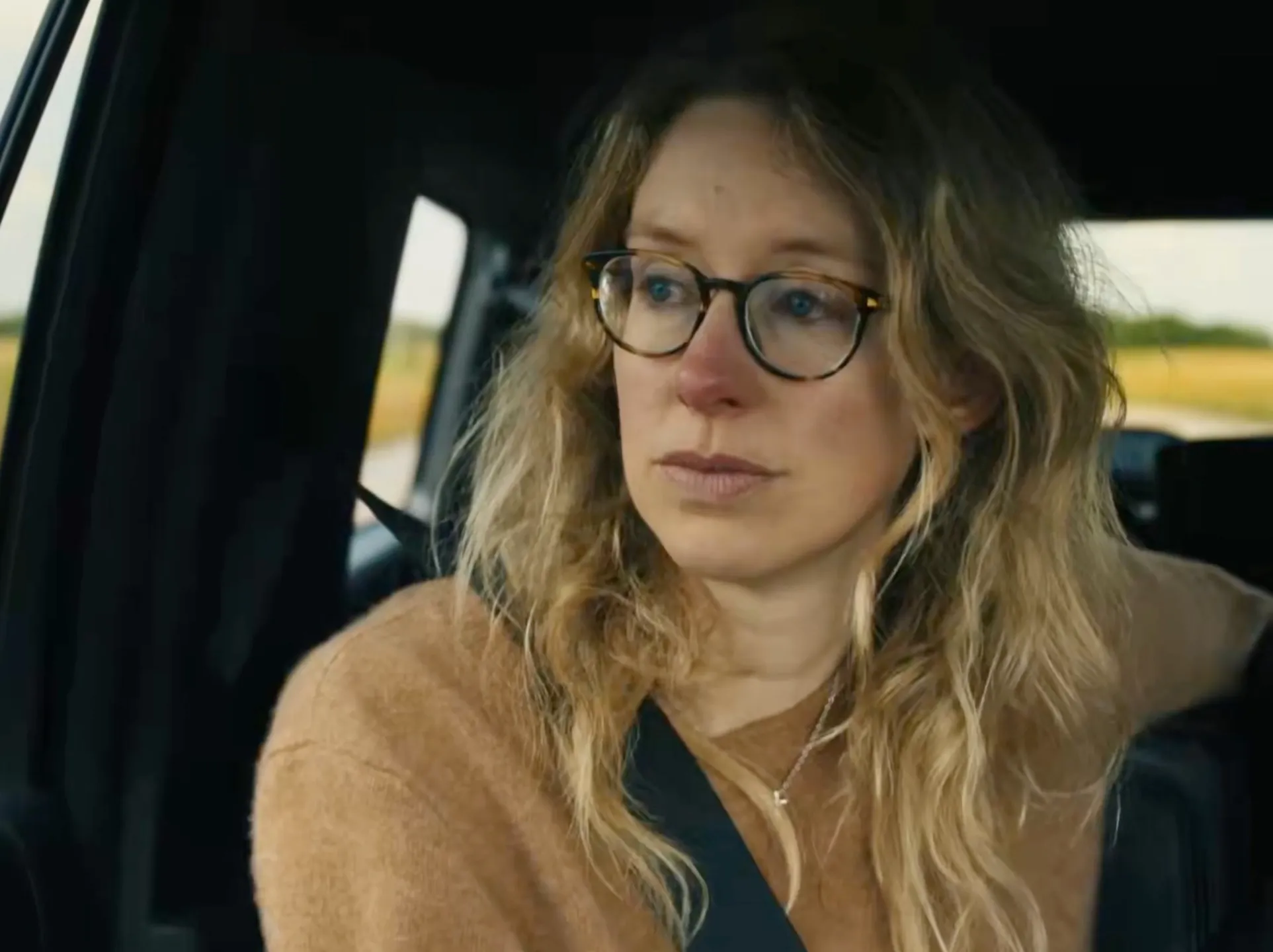 Elizabeth Holmes in a car, wearing glasses and a tan sweater, looking out the window with a pensive expression.