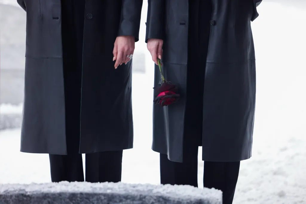 The bottom half of two individuals wearing black coats and trousers, one holding a black rose with red petals, standing in front of what appears to be a tombstone or a snow-covered surface.