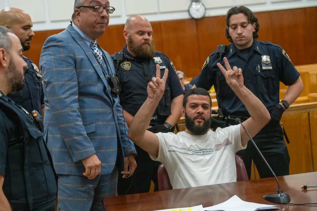 Yehia Amin making peace signs in court with police officers behind him.