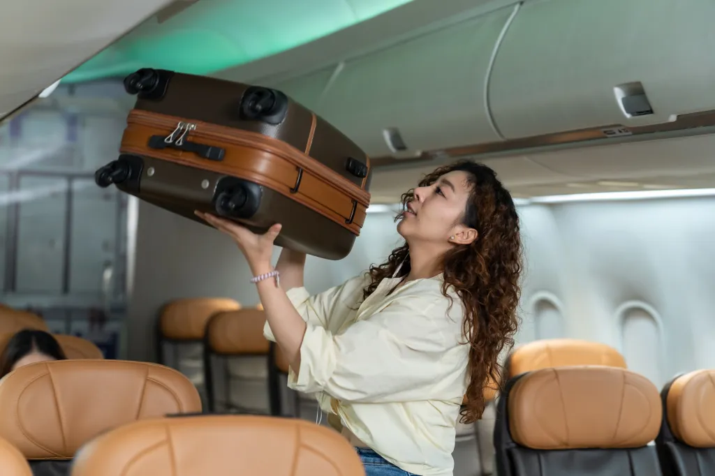 A young Asian woman lifts a suitcase toward an overhead compartment inside a commercial aircraft cabin on Saturday, Sept. 5, 2026.