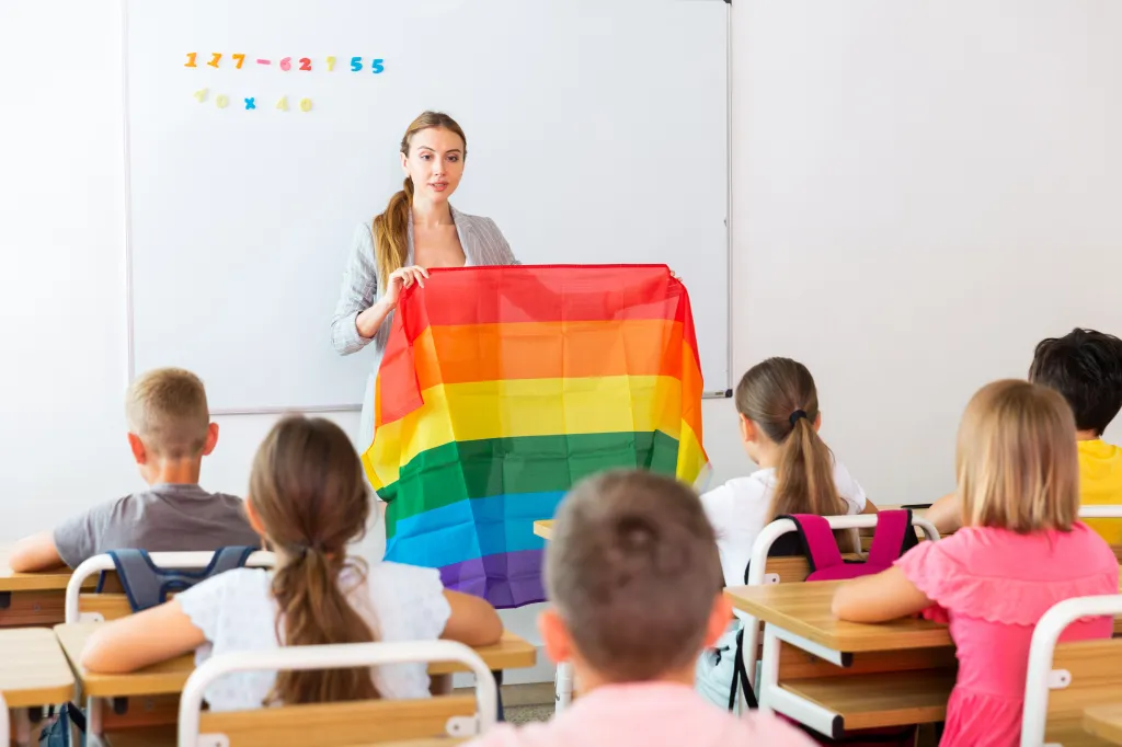 A female teacher discusses LGBT social movements with preteen children, holding a rainbow flag in a classroom.