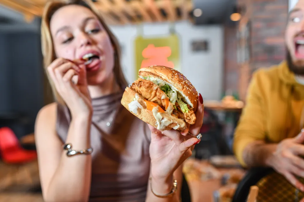 A young woman holding a crispy burger while reaching for a fry.