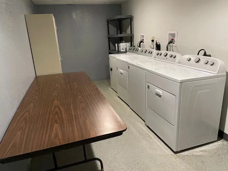 A laundry room with three washers and three dryers, a long wooden table, and a tall gray locker.