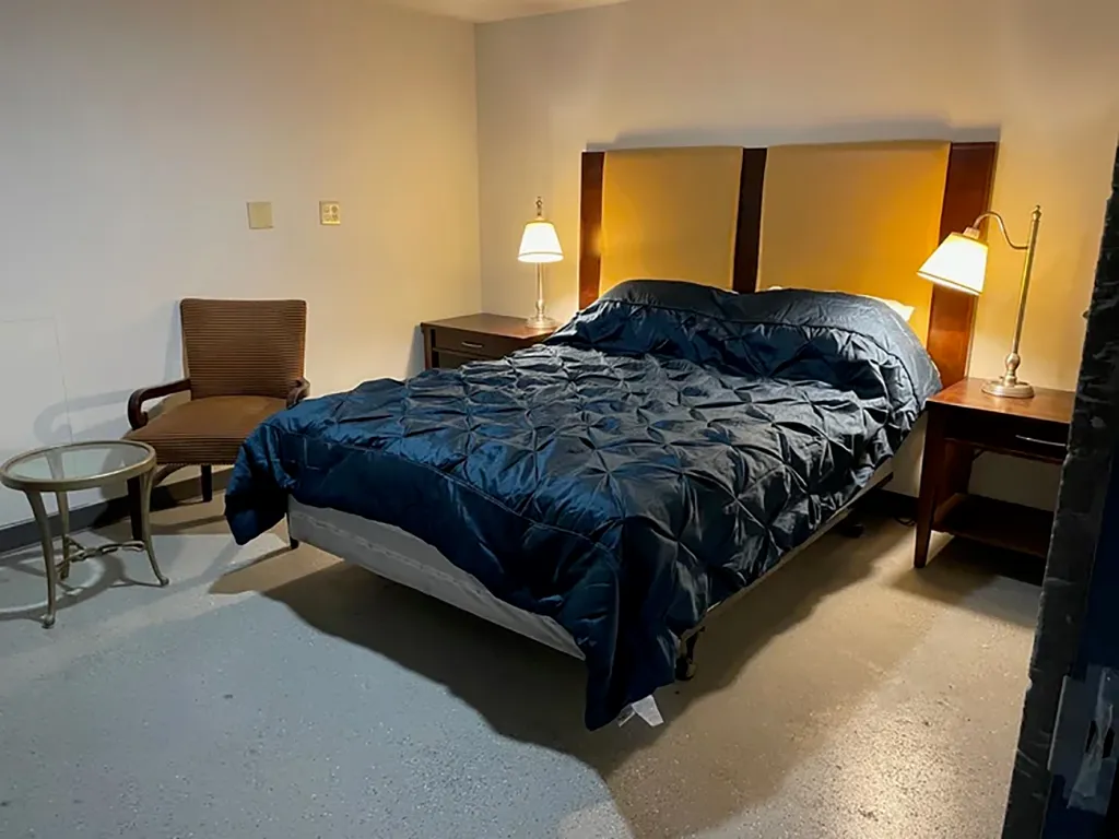 Bedroom in a bunker with a queen-sized bed, two side tables with lamps, and a wooden chair with a glass side table.