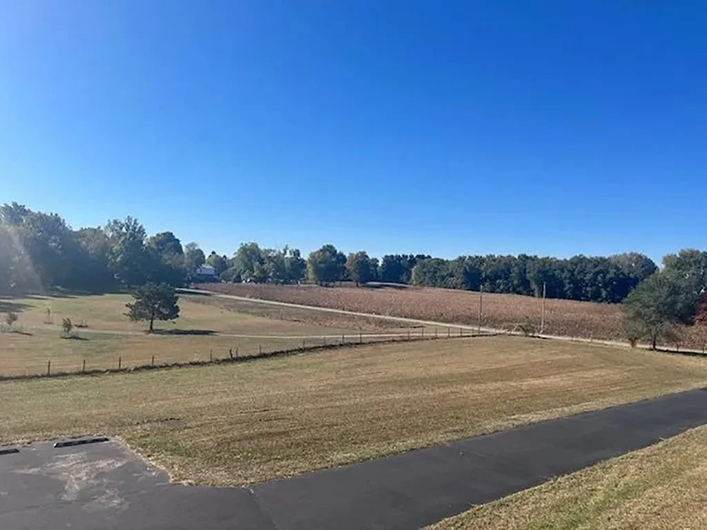 An empty field and a distant building surrounded by trees under a clear blue sky.