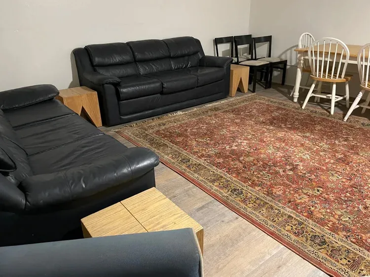 A room with two black leather couches, a red patterned rug, a dining table with chairs, and wooden side tables.