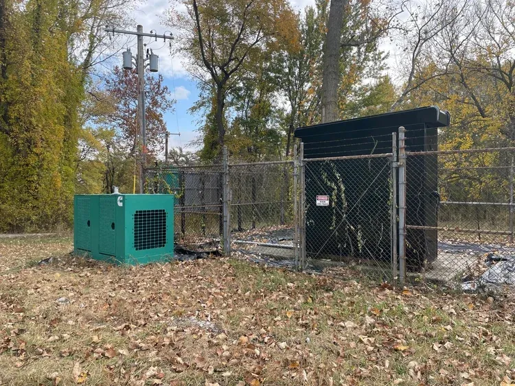 A green power generator next to a fenced-off electrical box, with trees and dead leaves in the background.