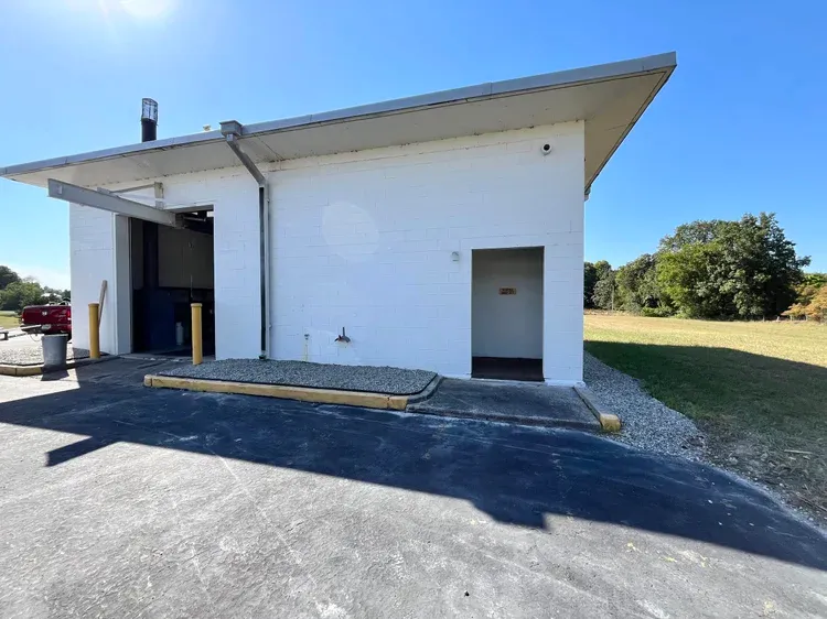A white brick building with a garage door and an open door on the side, set against a bright blue sky and green landscape.