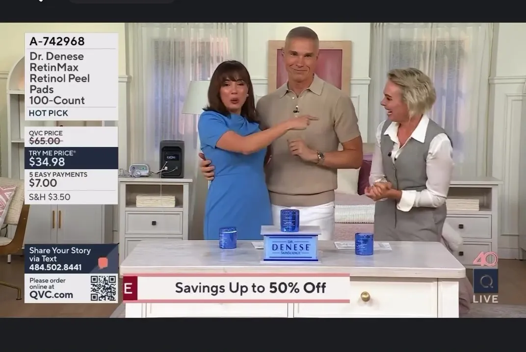 A QVC host in a blue dress points at her male co-host, while another female host laughs in the background.