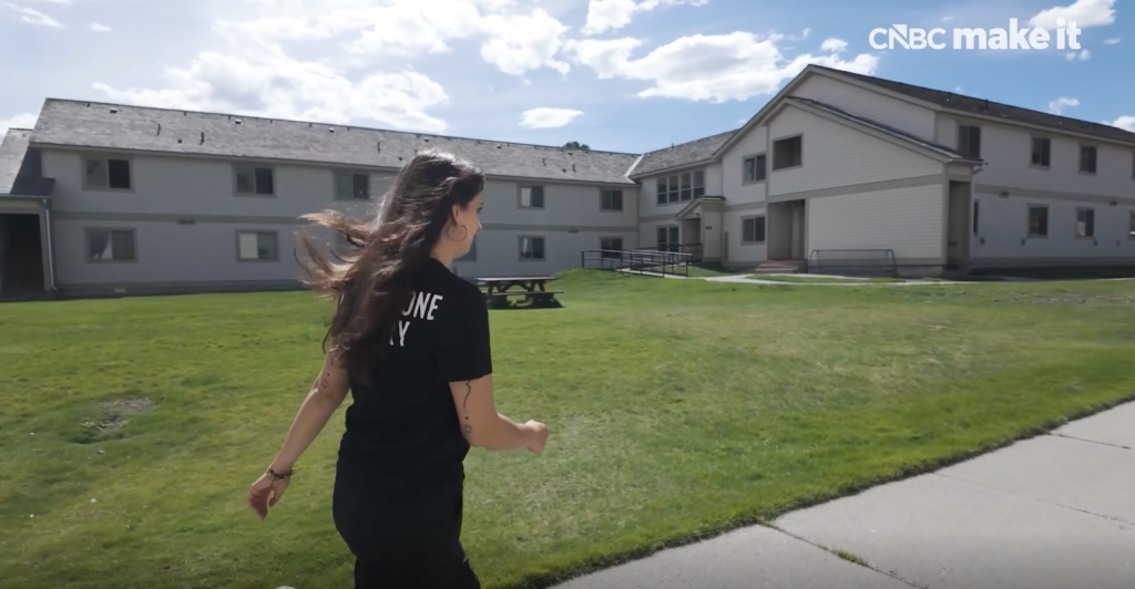 Zoe De La Paz walks across a grassy lawn with residential buildings in the background.