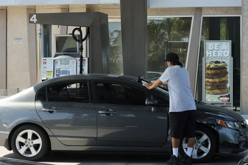 A man fills his car with gas next to a gas station convenience store advertising Zooies Cookies.