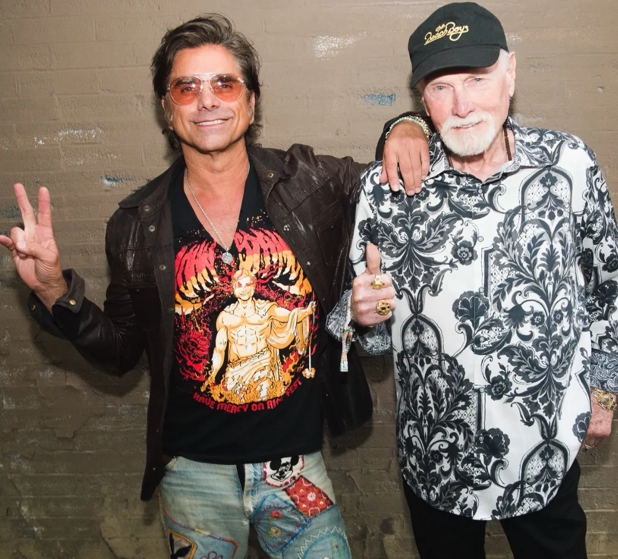 John Stamos and Mike Love standing side by side, with Stamos making a peace sign and Love giving a thumbs-up.