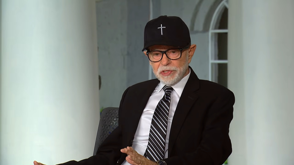 Jim Bakker wearing a black baseball cap with a white cross, glasses, a suit, and a striped tie.