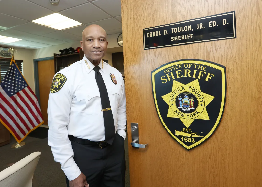 Suffolk County Sheriff Errol D. Toulon Jr. stands in his office next to a door featuring his nameplate and department insignia.