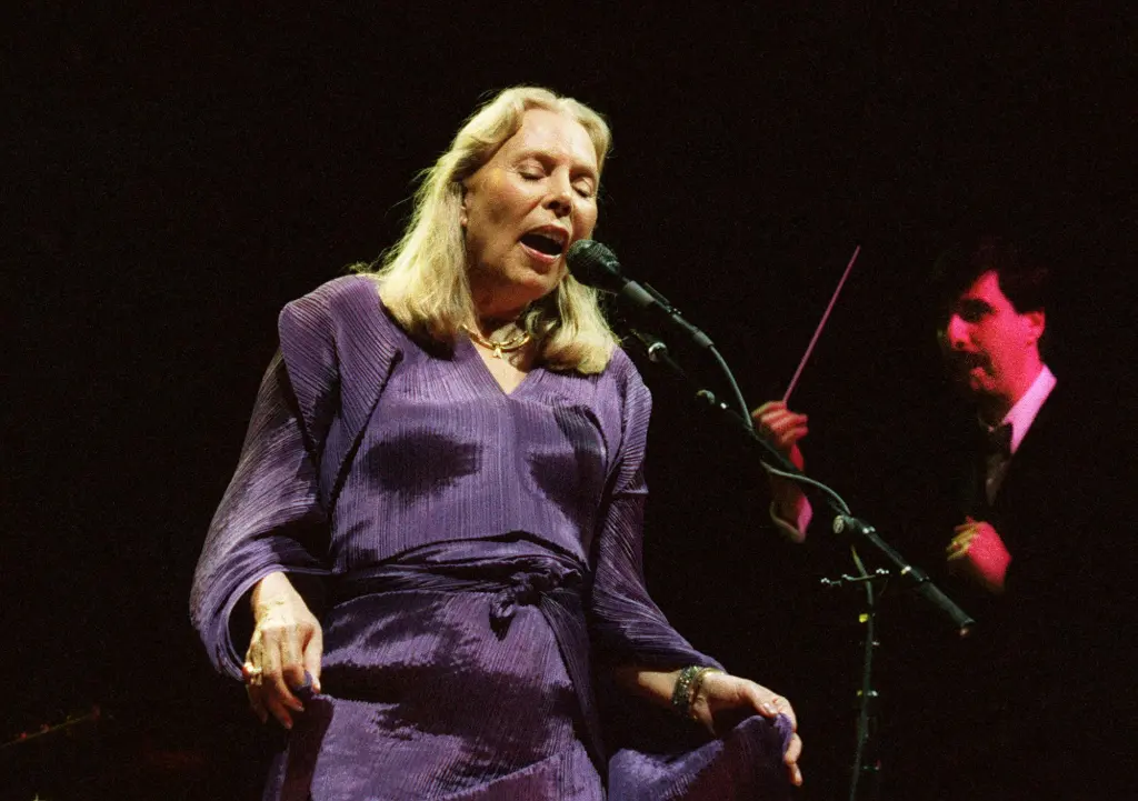 Joni Mitchell singing into a microphone on stage at the Madison Square Garden Theater.