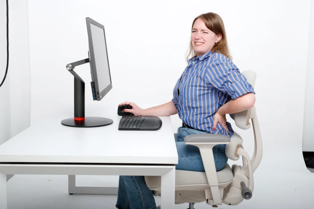 A woman sitting at a desk with a computer grimacing in pain while holding her lower back.