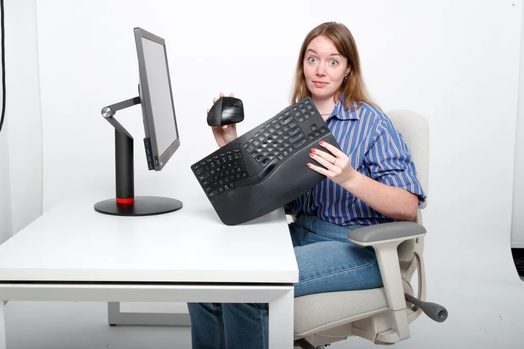 A woman at a desk holds up an ergonomic mouse and keyboard, looking skeptical.