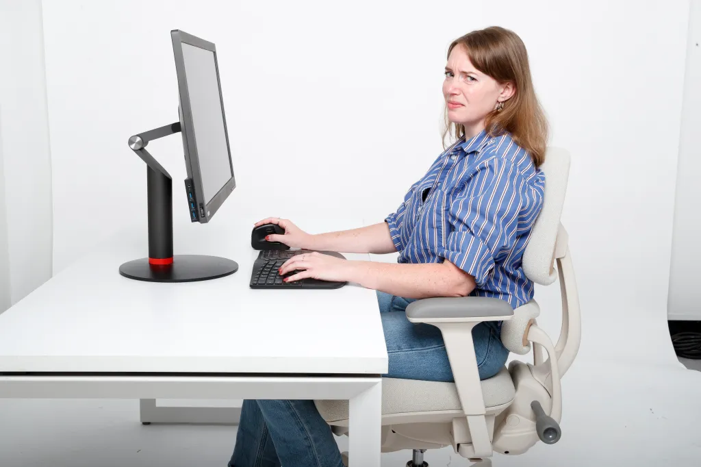 A woman sitting uncomfortably at a desk with a computer, demonstrating poor ergonomics.