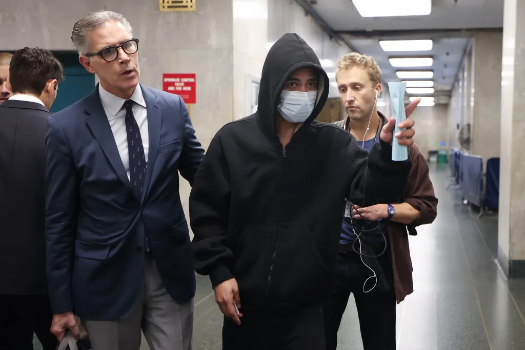 Samuel Reveron, wearing a black hoodie and face mask, is escorted through a courthouse hallway by an attorney and another person.