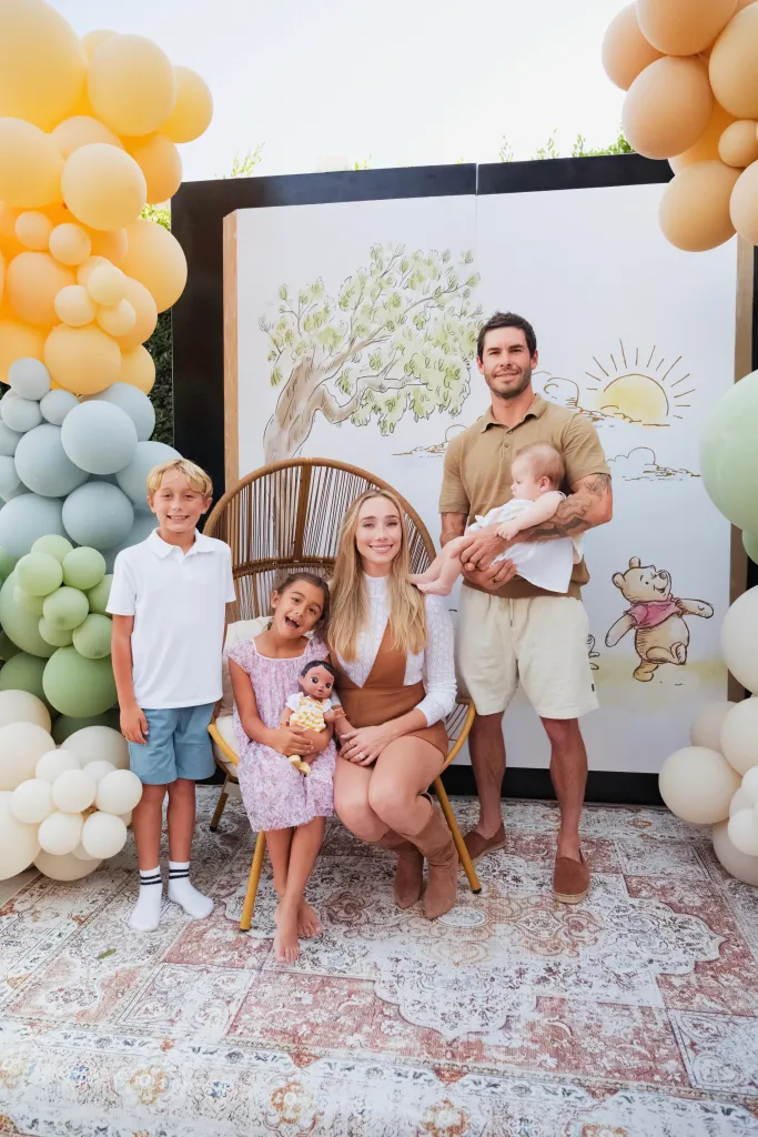 Nicole Williams English and her family pose in front of a Winnie the Pooh backdrop, surrounded by pastel balloons.
