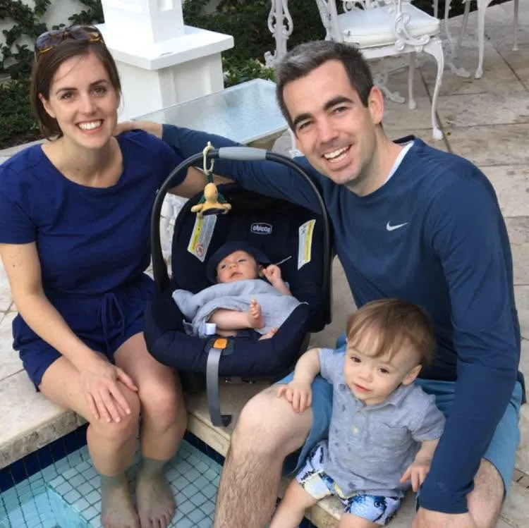 A family of four outdoors: a woman in a blue dress, a man in a blue long-sleeved shirt with a toddler on his lap, and an infant in a car seat between them.