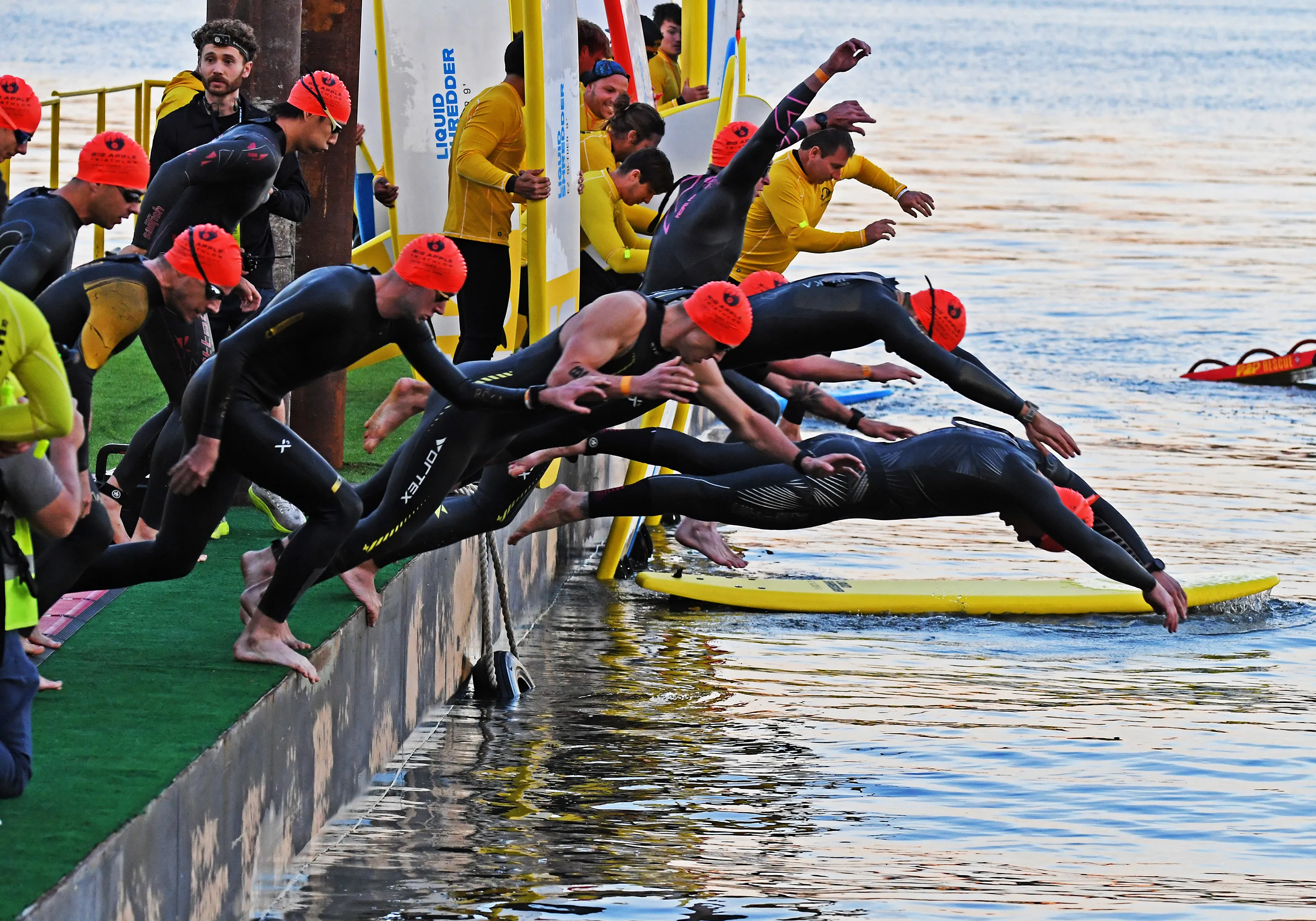 Competitors in wetsuits and orange swim caps leap from a dock into the Hudson River at the start of a triathlon.
