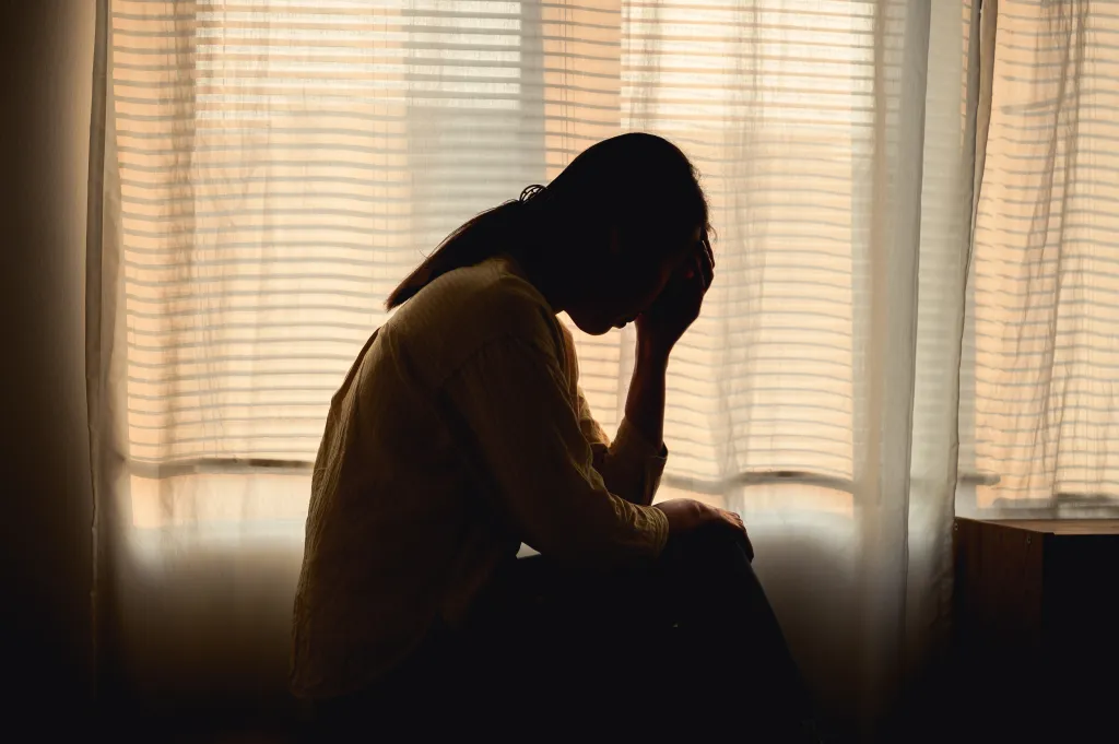 Silhouette of a depressed woman sitting by a window, holding her head in her hands.