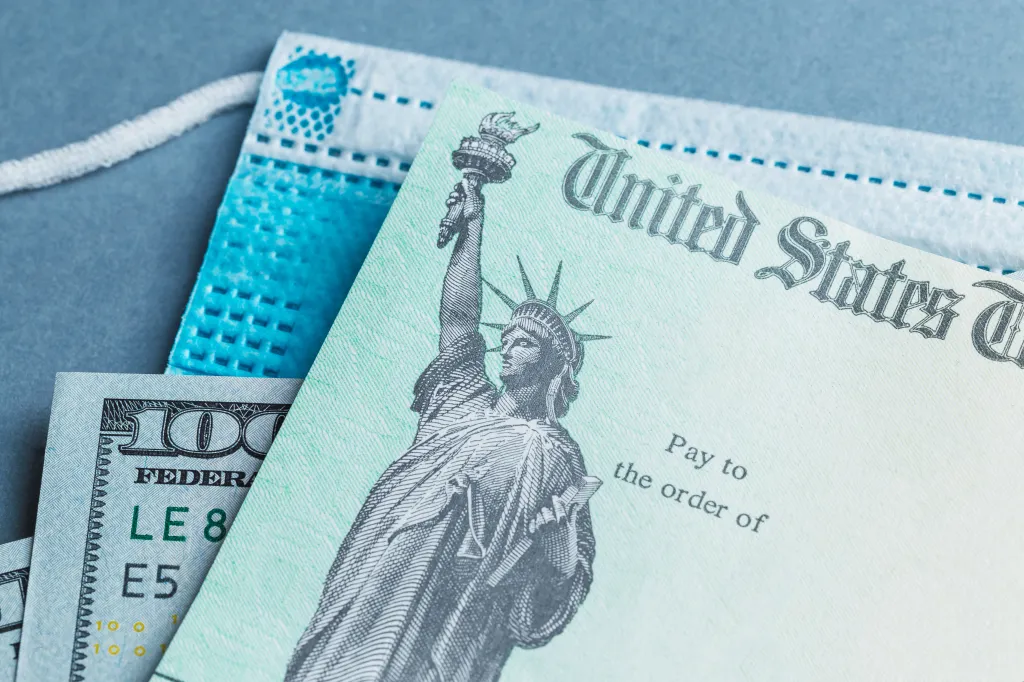 A US Treasury stimulus check sits atop a blue face mask, alongside US hundred-dollar bills.