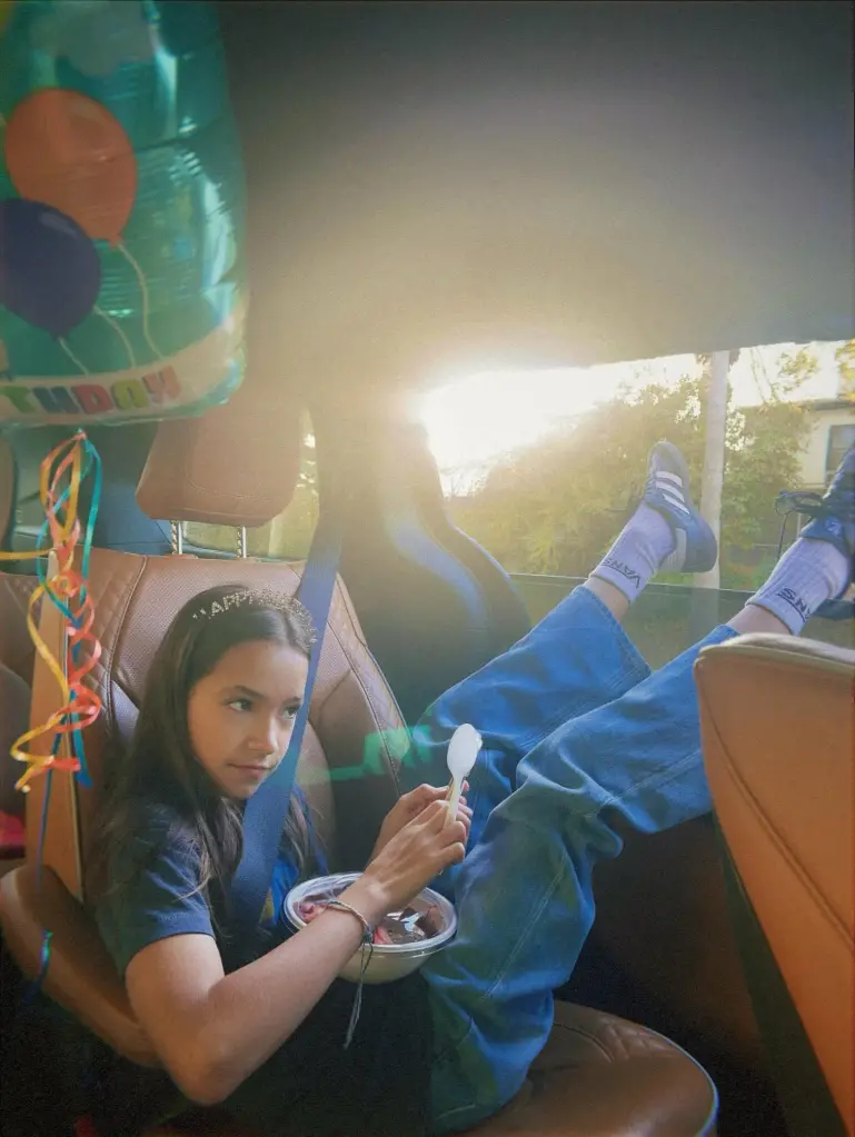 A young girl wearing a birthday headband sits in the back of a car with her feet propped up on the seat, holding a bowl of food.
