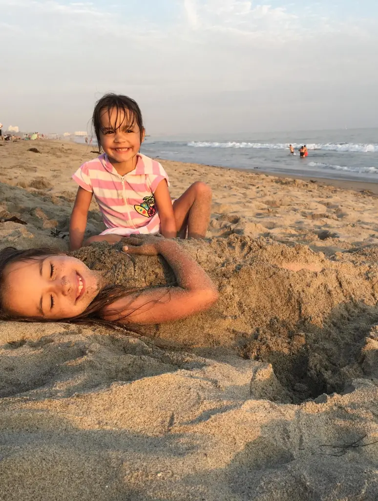 Two little girls play on the beach, one buried in the sand and smiling while the other crouches beside her.