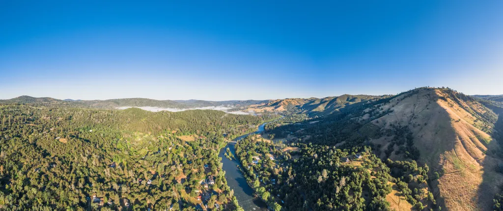 Aerial panoramic view of Coloma, California, featuring a winding river flowing through a forested valley surrounded by green hills under a clear blue sky.