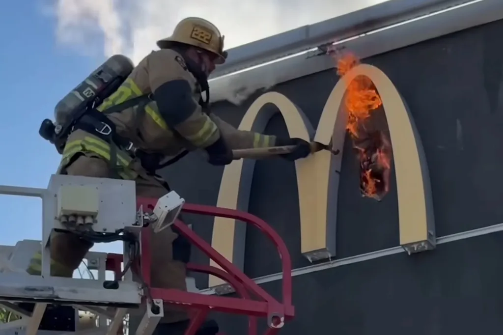 A firefighter on a platform uses an axe to smash a burning McDonald's sign.