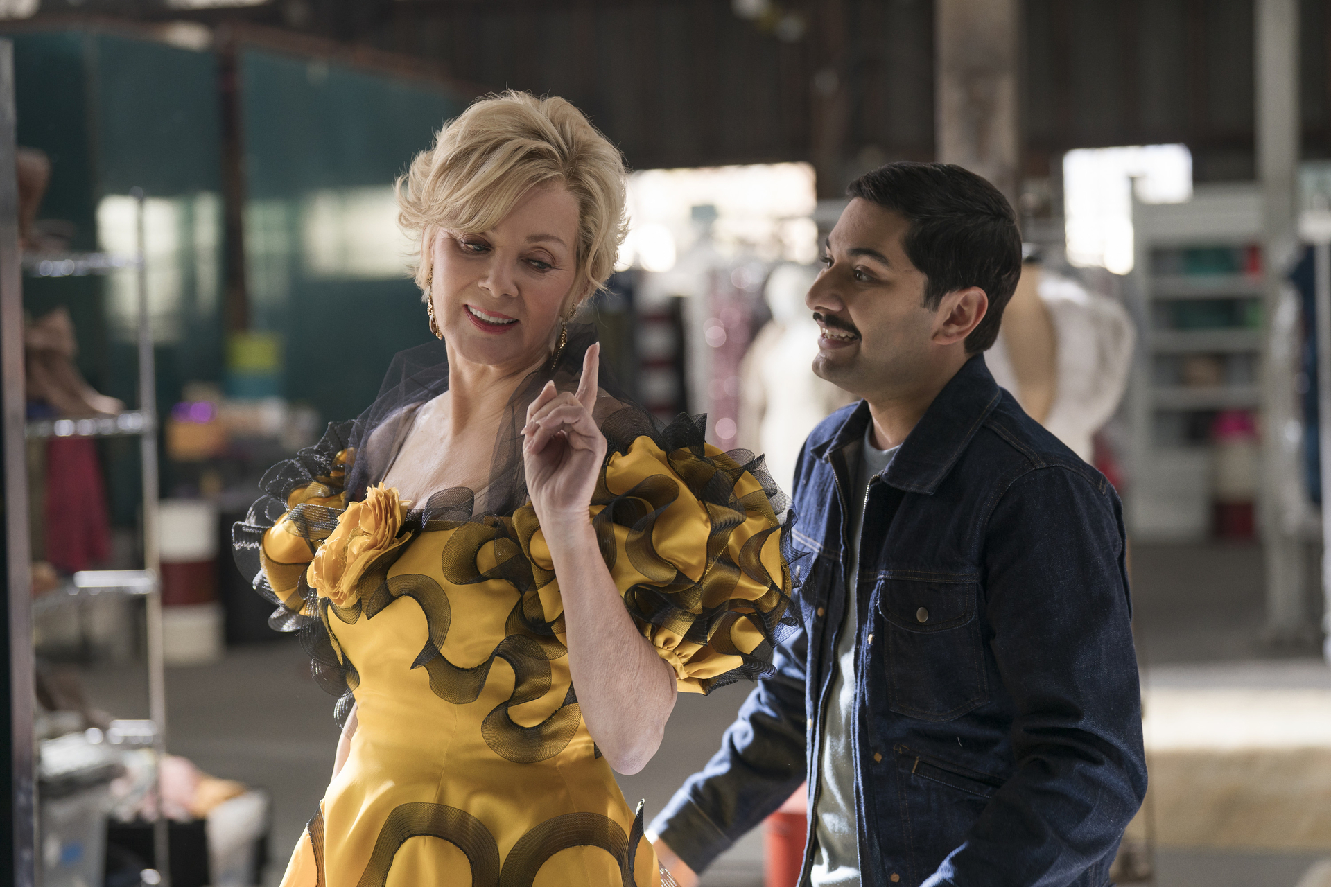 Jean Smart wearing a yellow dress with black ruffles, gesturing while a man in a denim jacket smiles at her.