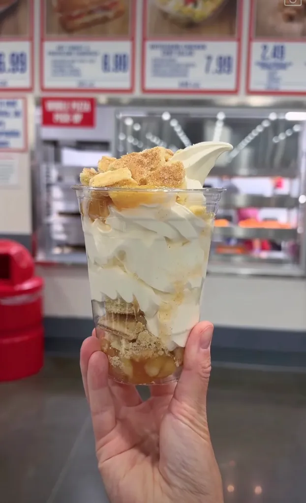 A hand holding a clear plastic cup filled with soft serve ice cream, topped with apple pie filling and pie crust pieces.