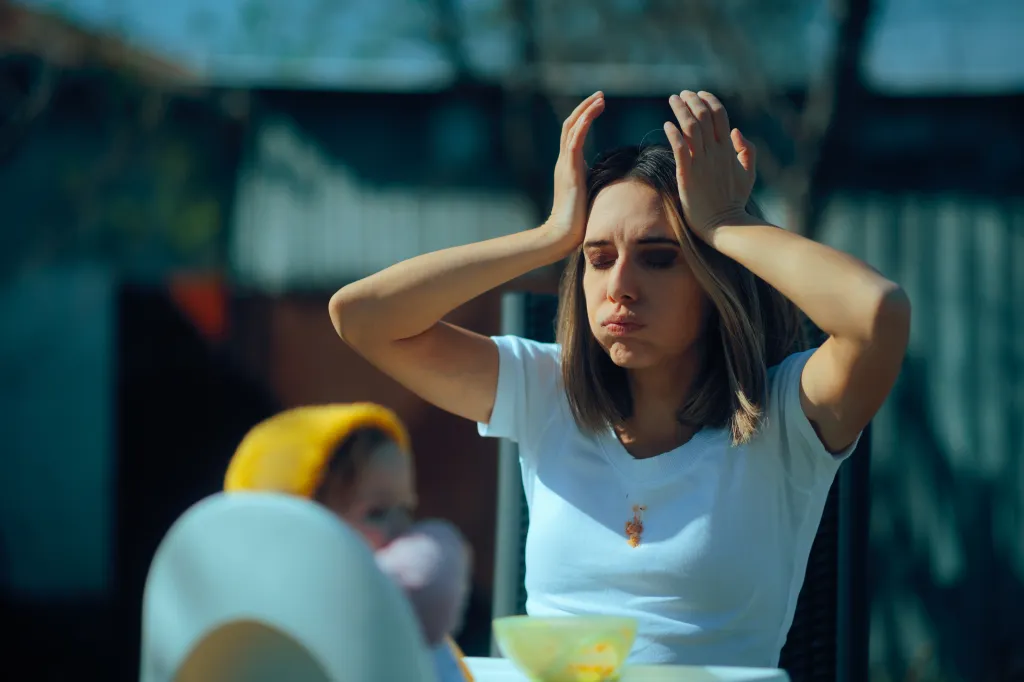 An overwhelmed mother looking stressed out with her hands on her head while trying to feed her baby.