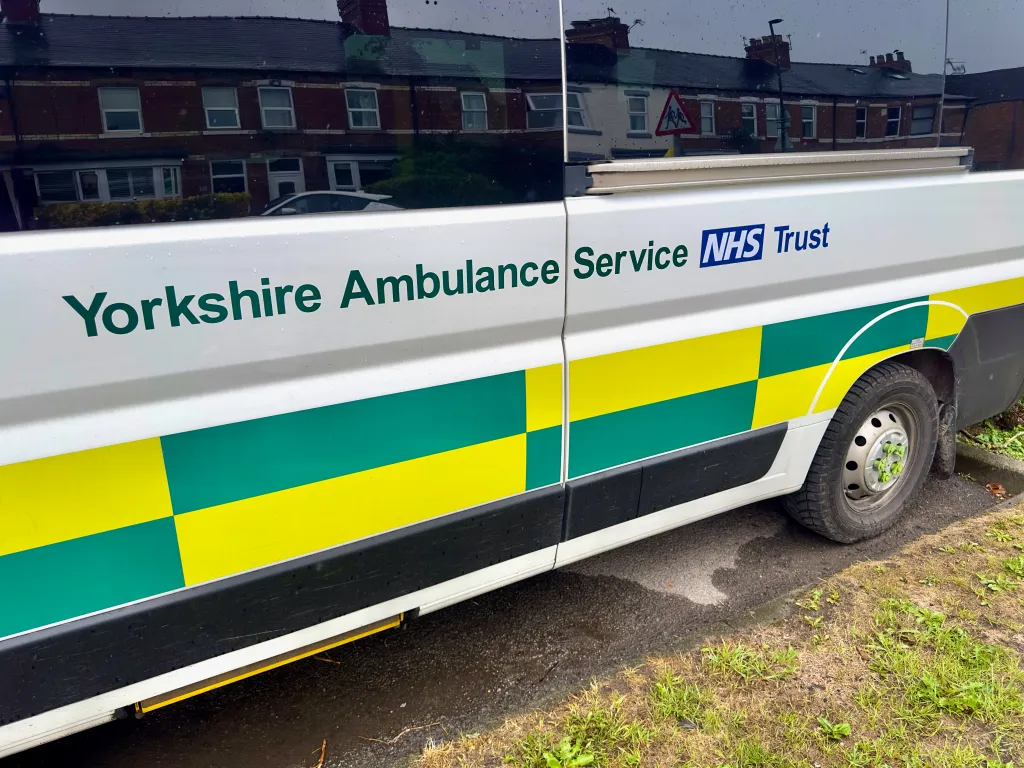 Side view of a white Yorkshire Ambulance Service van with green and yellow Battenberg markings and the NHS Trust logo.
