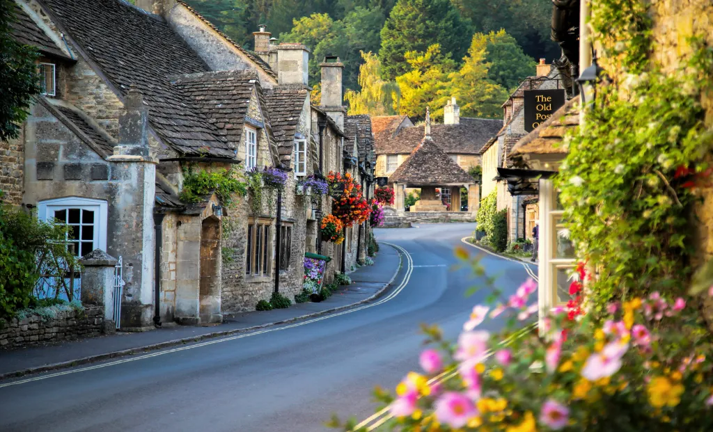 A street in Castle Combe, England, lined with historic stone cottages and colorful hanging flower baskets.