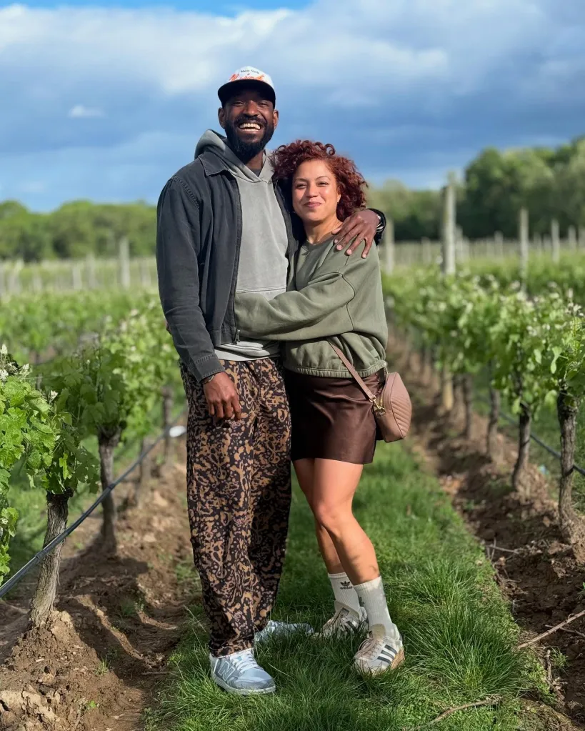 A smiling man and woman pose embracing each other in a vineyard under a cloudy sky.