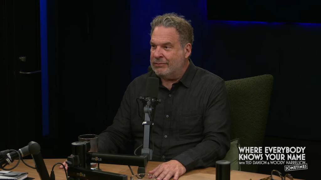 Comedian and actor Jeff Garlin sitting at a microphone in a dark studio.