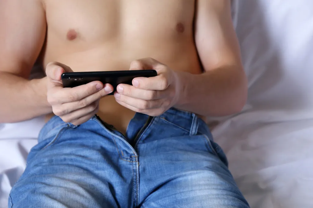 A shirtless man holding a smartphone in landscape orientation while lying in bed in unzipped jeans.
