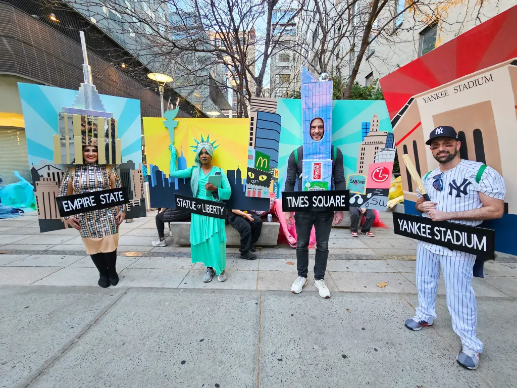 Four people in New York City landmark costumes: the Empire State Building, the Statue of Liberty, Times Square, and Yankee Stadium.