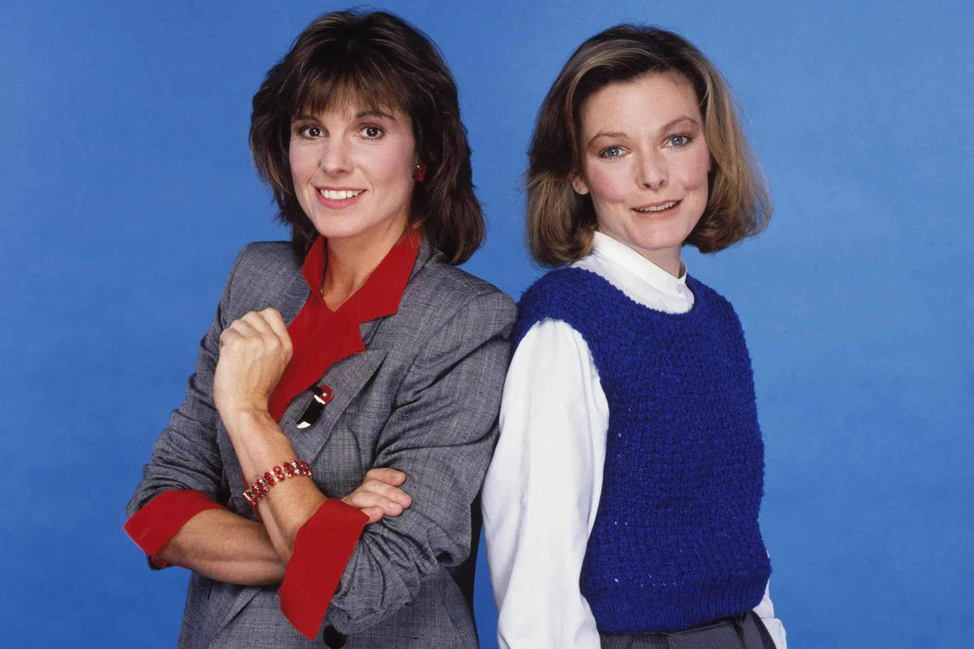Two women, Penny Marshall and Cindy Williams, stand back-to-back against a blue background, both smiling at the camera.