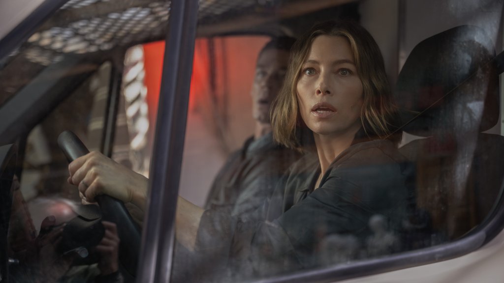 Jessica Biel driving a van with a worried expression while looking ahead, with a male passenger visible in the background.