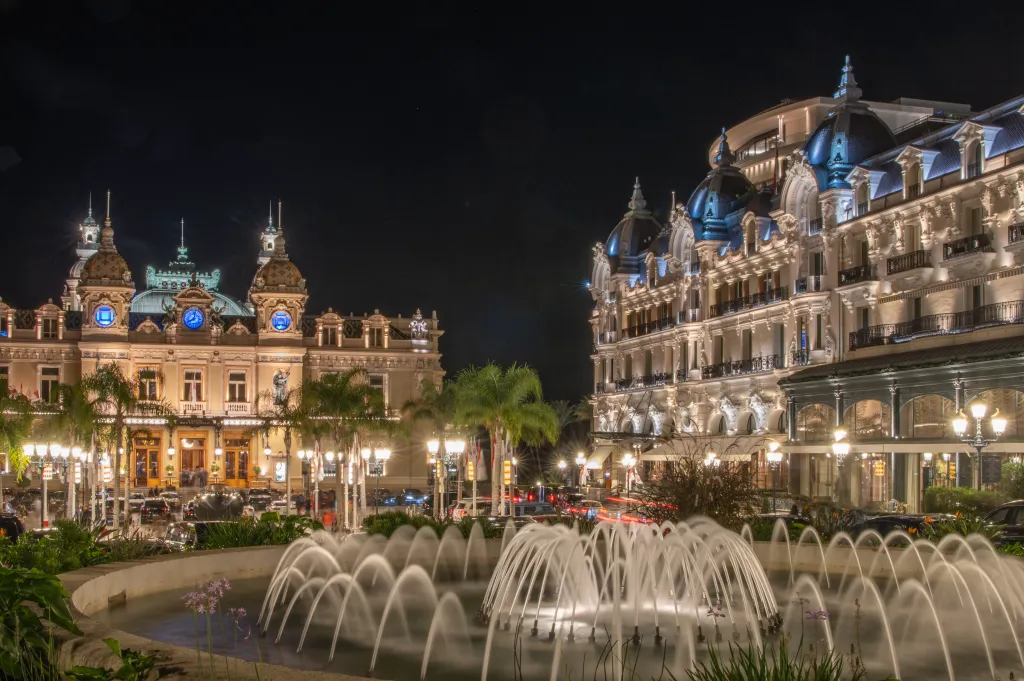 A night view of the illuminated Casino de Monte-Carlo and Hotel de Paris in Monaco, with a brightly lit fountain in the foreground.