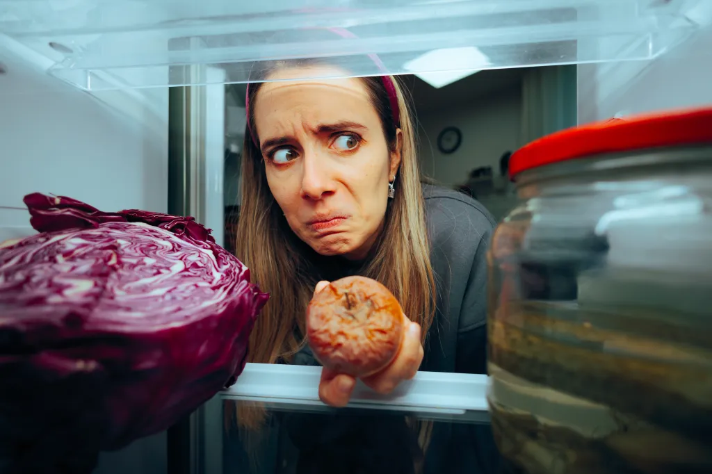 A woman looks into a refrigerator with a disgusted and worried expression, holding a rotten apple in her hand.