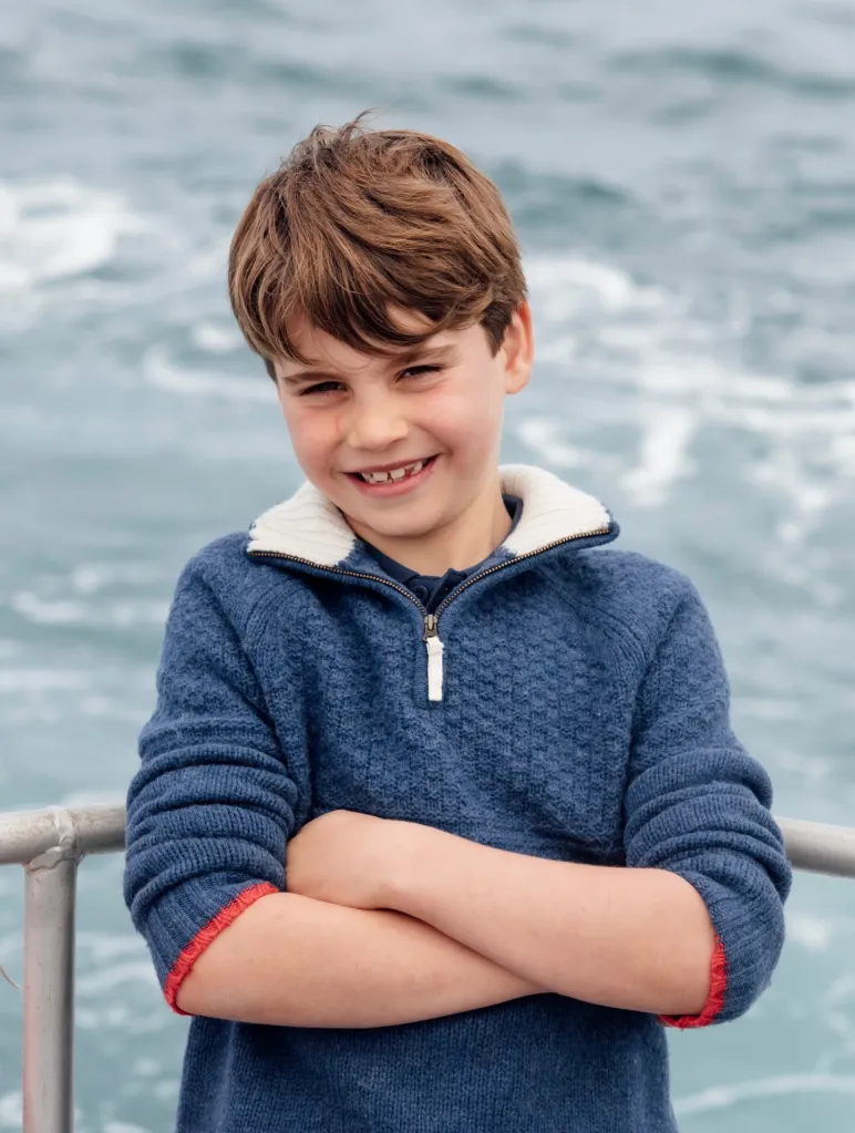 Prince Louis smiling with arms crossed against a backdrop of water.
