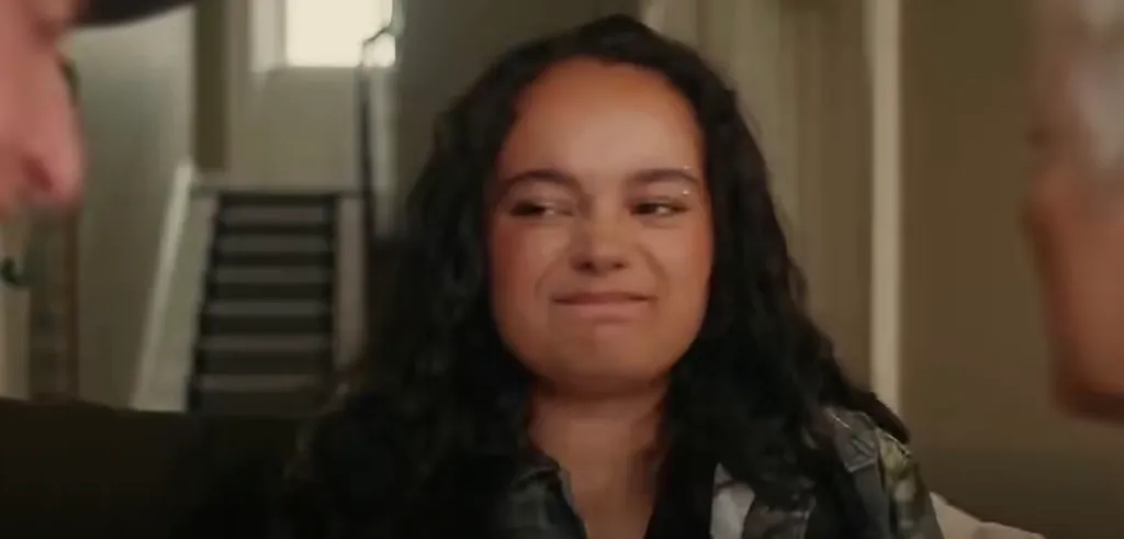A young woman with dark curly hair looks uncomfortable between two adults in a scene from the public service announcement.